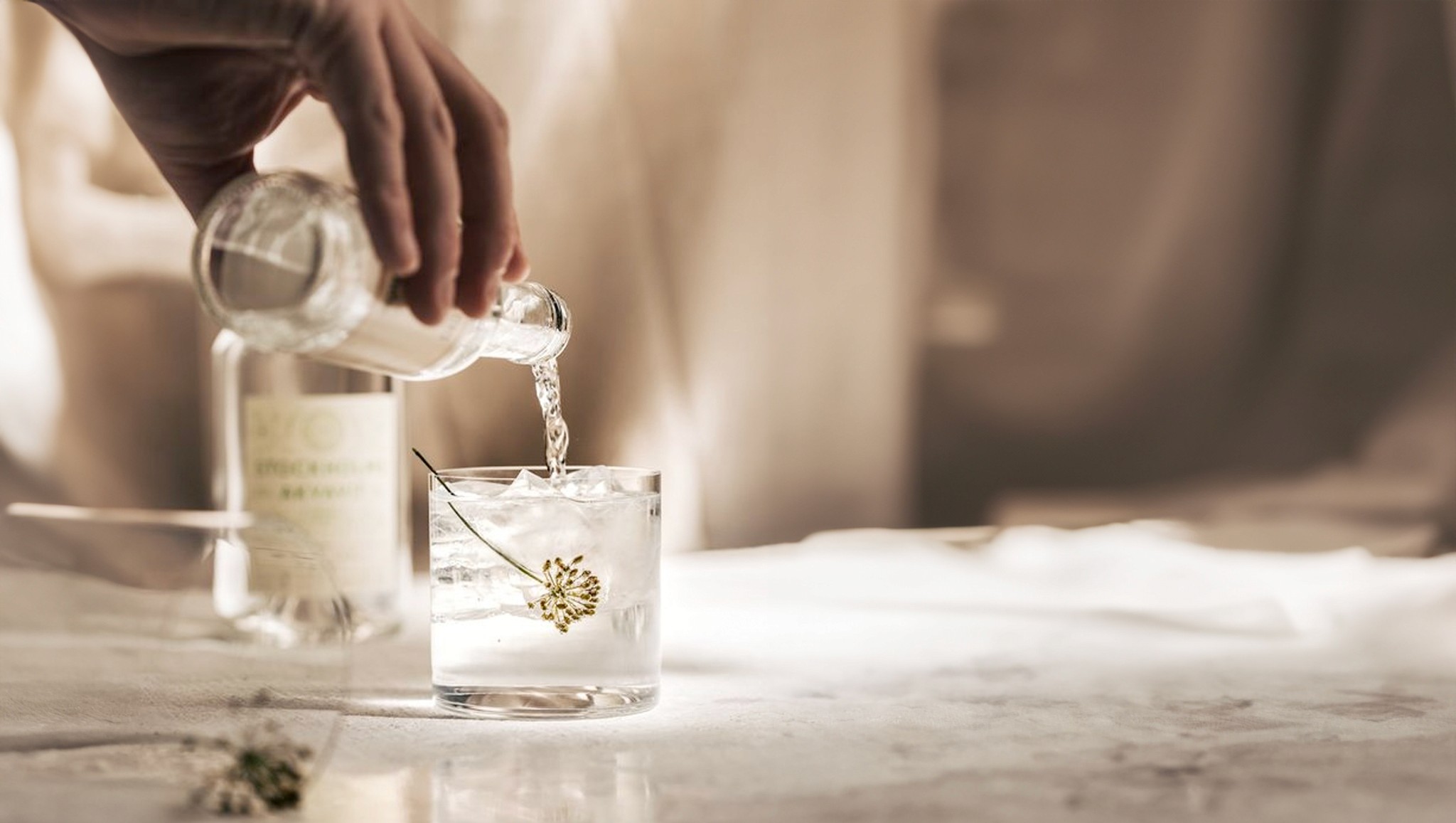 Eine Hand gießt einen klaren Drink über Eis in ein Glas, daneben getrocknete Kräuter.